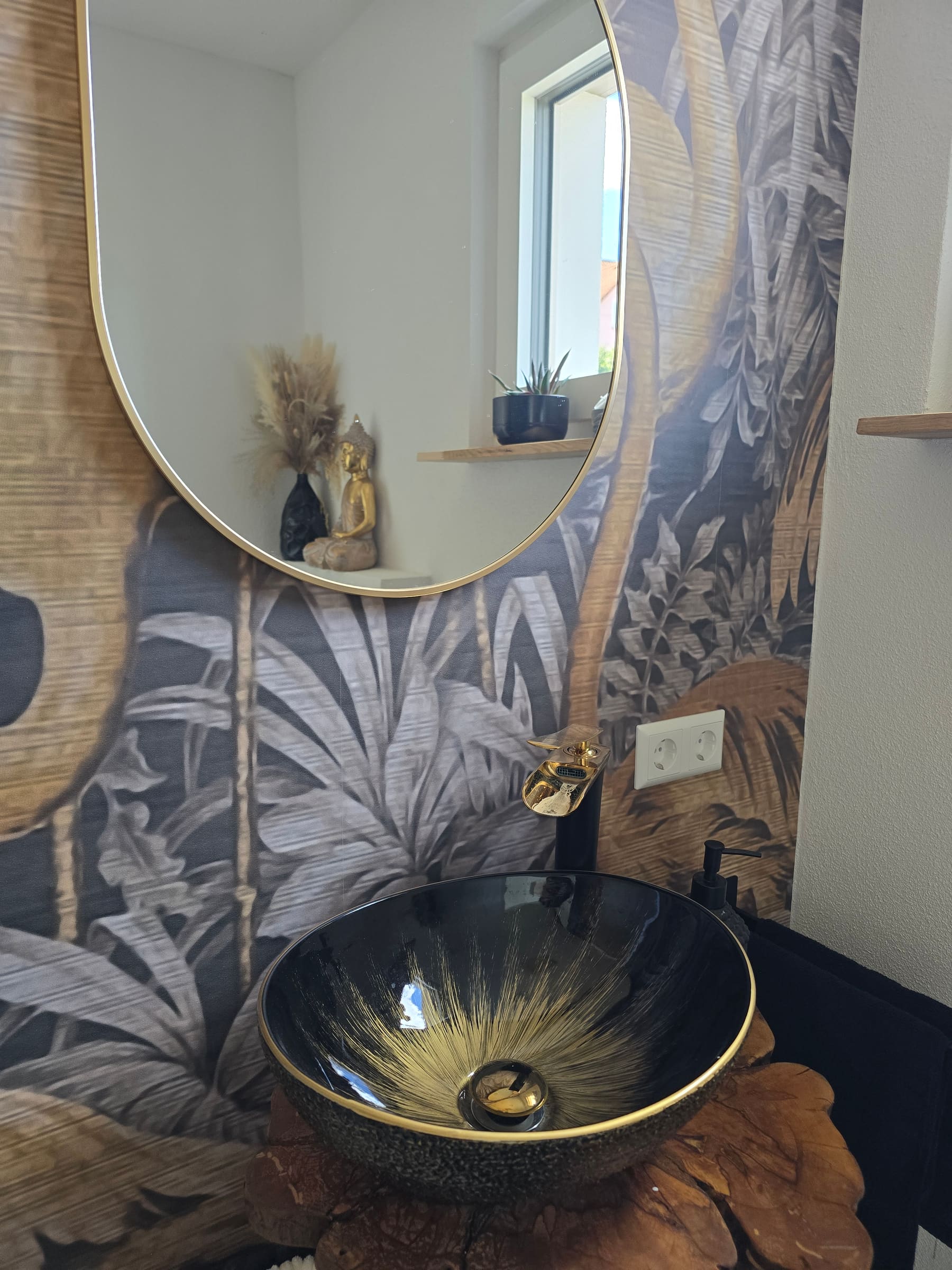 Schwarzes Waschbecken mit goldfarbenen Details vor einer botanischen Wand